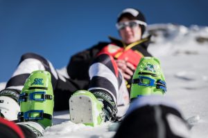 Noemi im HIntergrund unscharf im Schnee. Im Vordergrund die grün leuchtenden Dalbello Skischuhe von Ihr und Ihrem Guide