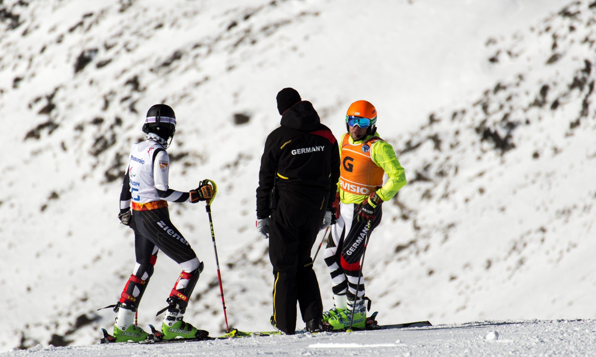 Auf dem Bild sind die Skifahrer Noemi und Lucien mit Ihrem Trainer zu sehen. SIe betrachten den Hang und besprechen sich.